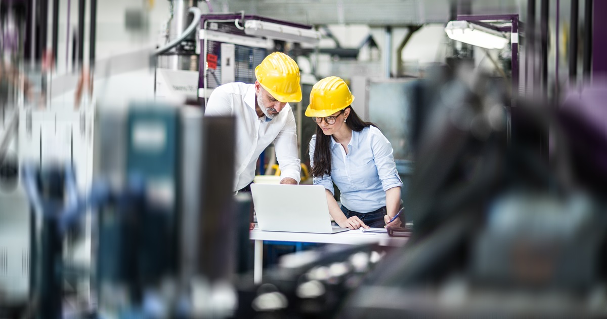 Two project managers standing in modern industrial factory, looking at laptop screen. Manufacturing facility with robotics, robotic arms and automation. Storing products and materials in warehouse.
